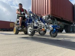 Man in a safety vest rides a small motorized tricycle as a large shipping container is loaded onto a trailer in a cargo yard.