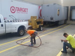 Two workers in a loading dock area: one in an orange shirt handling a long hose near a white pickup and trailers (Target and UPS).