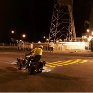 Nighttime industrial area with a man in a yellow shirt and helmet riding a small motorized bike across a yellow crosswalk.