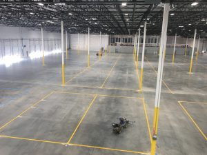 Interior of a large empty warehouse with yellow safety pillars, blue floor markings, and an exposed metal truss ceiling.
